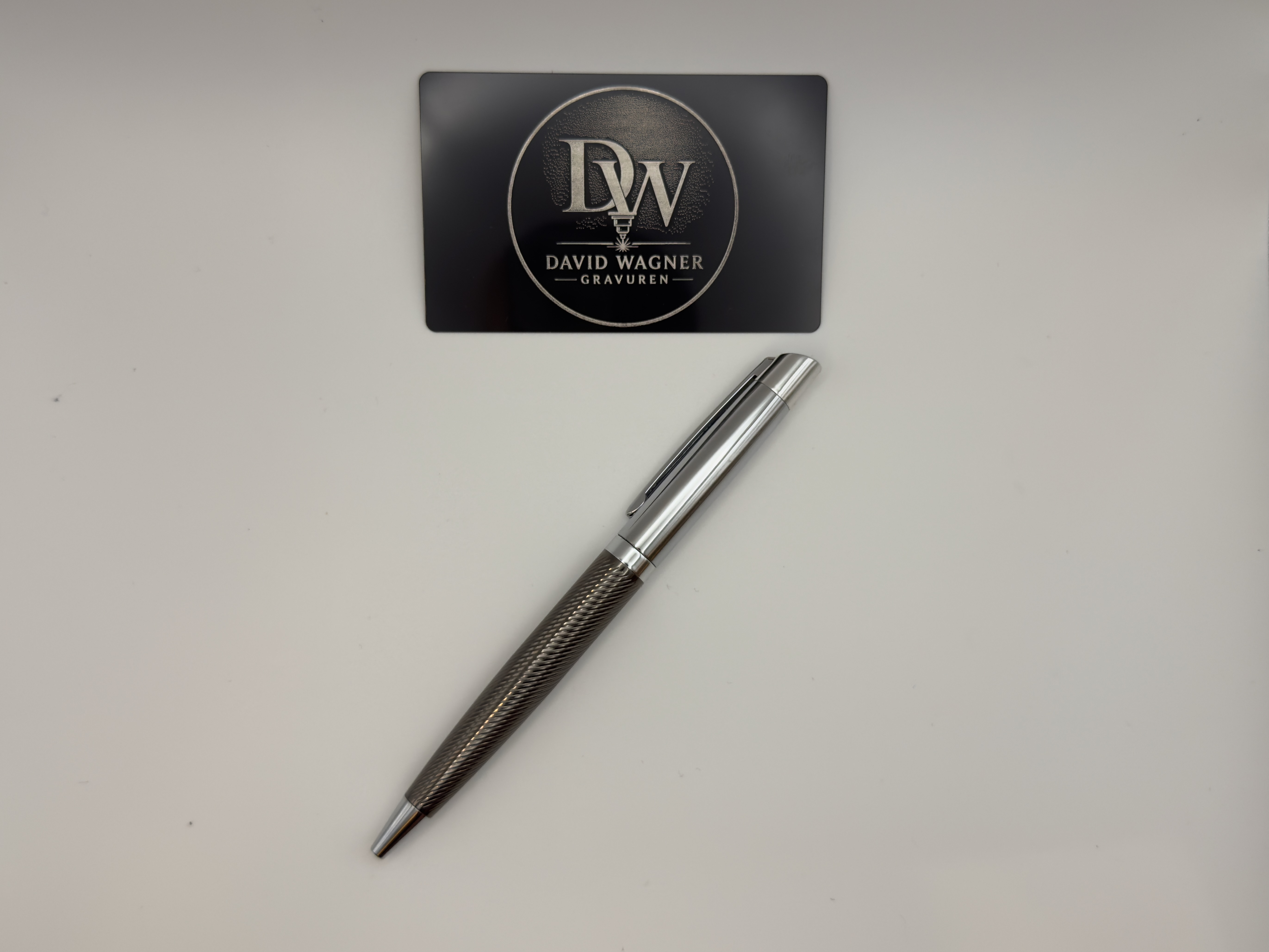 Schwarze Aluminium-Musterkarte mit lasergraviertem DW-Logo neben einem Edelstahl-Stift